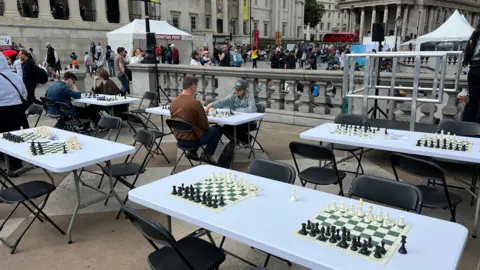 Five table with chess sets in Trafalgar Square with people standing around in the background 