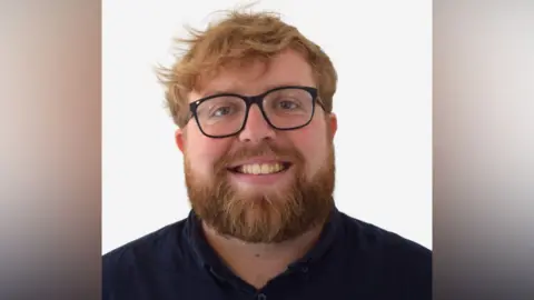 Luke Smout A photo of Luke Smout against a white backdrop. He smiles at the camera. He has light red hair and a thick red beard. He wears glasses and a dark navy blue shirt. 
