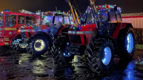 Suffolk Tractor Light Parade Two tractors and a fire truck covered in Christmas lights while parked up next to each other.