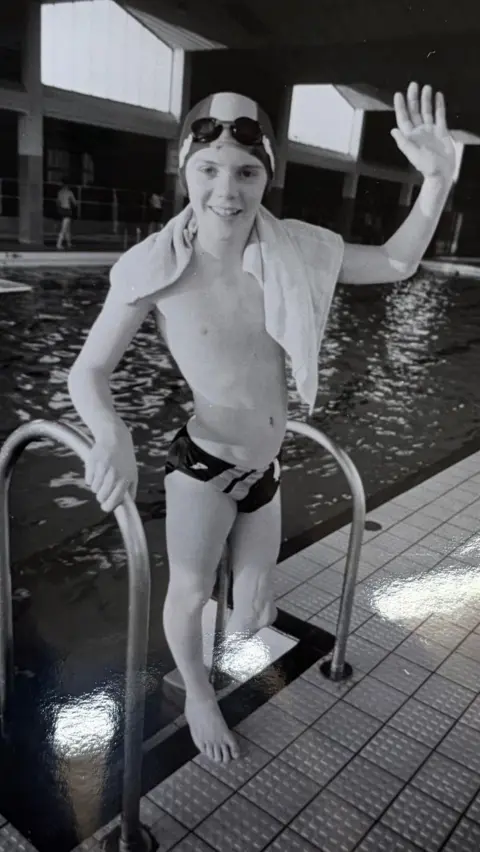 Mark Williams A black and white photo of Mark as a child, waving at the camera from the steps of a swimming pool. He is wearing swimming trunks and a swimming cap, and his left leg is amputated below the knee.