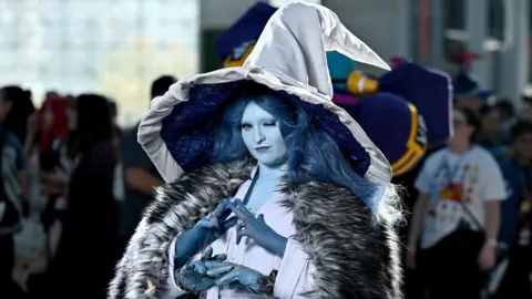 A fan of Elden Ring at last year's Comnic Con in New York dressed as Elden Ring's Ranni the Witch, in a grey gown, huge silver hat, fur cape and blue skin make-up, with an extra set of arms and hands and long, blue hair