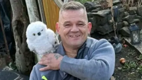 Family A man with brown hair and a blue top has a white owl on one shoulder. He is looking at the camera.
