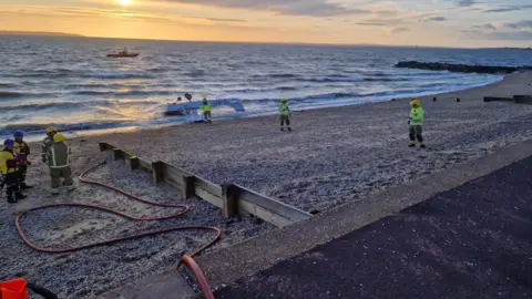 Hampshire & Isle of Wight Fire and Rescue Service A light aircraft is upside down in shallow water by the beach with emergency services personnel standing nearby