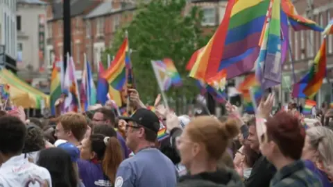 A crowd of people holding rainbow flags in a city street, standing in profile.