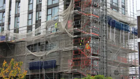 Builders work on a construction site in london. Three men can be seen climbing scaffolding while wearing fluorescent jackets and hard hats.