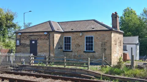 Friends of the S&DR Long and short brick building with windows and a chimney. In front of it is a fence and a railway line. Behind it is a white building. 