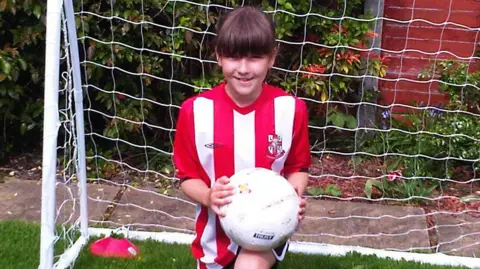 Alan Clark Olivia Clark, as a girl, smiling at the camera. She is kneeling and is holding a white football. She is wearing a red and white striped football shirt and is in front of a football goal.