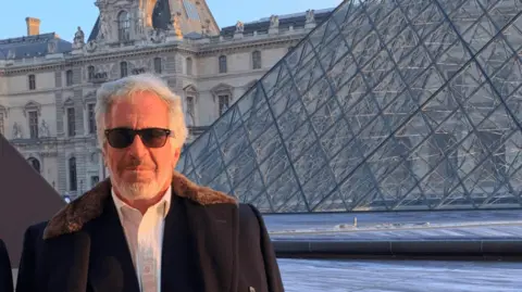 US Department of Justice Jeffrey Epstein, with a grey beard and wearing a fur-trimmed coat draped over his shoulders and sunglasses, stands in front of the Louvre and its glass pyramid.