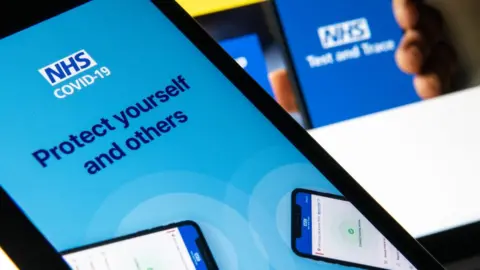 Getty Images The NHS Covid-19 app logo is seen on a smartphone, with branding from the website for test and trace behind it