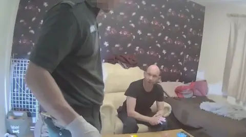PA Media A bald man in a black t-shirt and dark trousers sits on a cream armchair in a room with dark floral wallpaper behind and a single bed next to him with brown duvet and pink cushions. He is seen talking to a male paramedic, in dark green uniform, whose face can't be seen. A dog's bowl is on the left and a clothes airer.