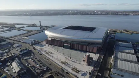 An aerial view of Everton Football Club's new Bramley-Moore Dock stadium. It is a large silver structure with a curved roof. The stadium is on the banks of the River Mersey.