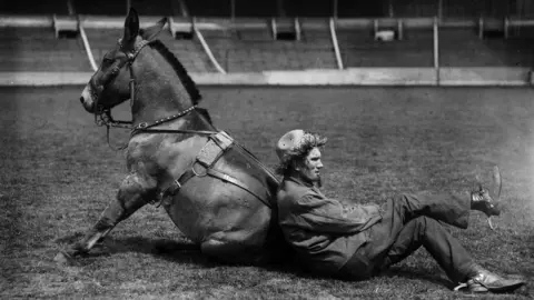 Getty Images Black and white image of a rodeo performer in costume leaning against a mule