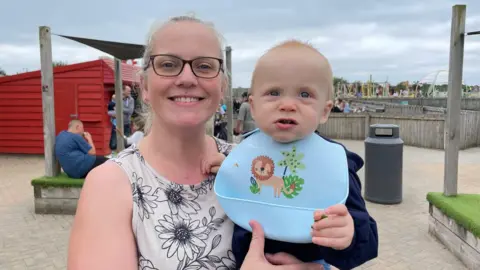 Michelle Birkenhead looks straight at the camera and smiles while holding a baby wearing a blue bib with a picture of a lion on it. She is smiling, wearing glasses and a  sleeveless white top with black flowers on it. She is outside and the sky behind her is overcast. 