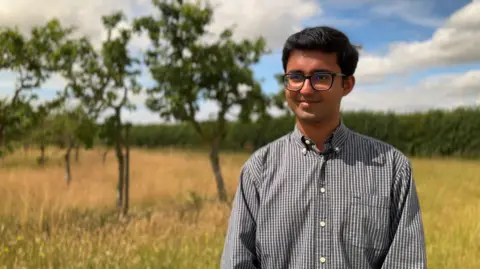 A man wearing glasses and a grey-coloured shirt with open collar. He is standing outdoors in an orchard and is smiling for the camera.