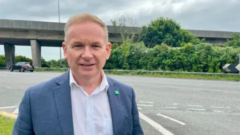 John Stephens, the cabinet member for transport, in a white shirt and blue blazer with a green Plymouth City Council badge
