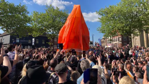 BBC/Ben Sidwell A view from the back of a large crowd standing outside in a square with phones raised ready to take a picture. A red sheet covers a statue at the centre of the photo. Buildings surround the square and the sky is blue.