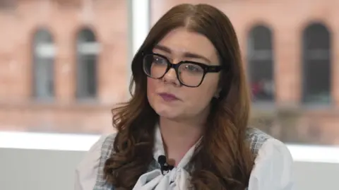Jennifer Lilly is a young woman with long red-brown hair, large plastic spectacles and a white blouse. She is in an office with a red sandstone building visible through the window.