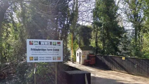 A view of the front entrance of a farm camp for agricultural workers. There is a sign with the name of the company - WMS - and the site name - Fridaybridge Farm Camp. There is a small road going across what looks like a bridge into the camp and a small wooden shed with pallets stacked against one side