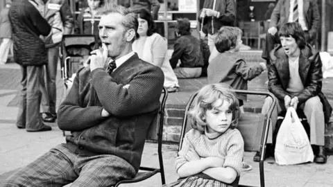 Heidi Alexander Man and young girl sitting on chairs at the market