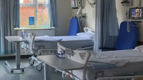 Part of a hospital ward, showing two new, empty beds with medical equipment.