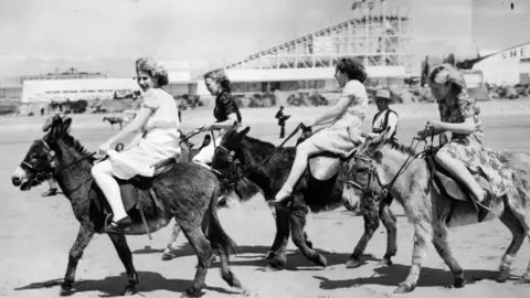 Getty Images A black and white image of people riding donkeys on Porthcawl beach with the fairground rides seen in the background.