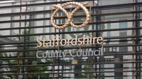 Image of Staffordshire County Council logo at the front of the authority's main building in Stafford. It includes the staffordshire knot, and the wording of staffordshire in a gold font. 