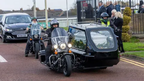 Mr Dingwall's coffin being carried in a black hearse sidecar attached to a motorbike. His brother, Robert is riding the motorcycle. Another biker, with a large white beard is riding behind. People are standing at the side of the road to pay their respects.