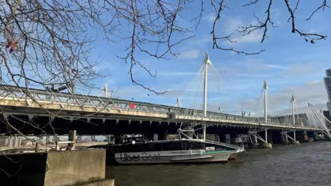 Network Rail A landscape shot of the Victorian-style bridge from Victoria embankment with the River Thames running beneath it and bright blue sky. Outward leaning pylons run along each side and there is an Uber Boat in motion beneath the bridge. 