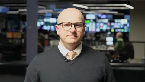 A close-up photo of Carwyn Donovan, a bald man with glasses, V-neck sweater and a gold tie.