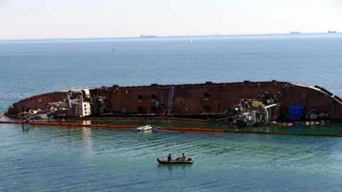 Getty Images The Delfi oil tanker, pictured on its side in July 2021 as workers try to use absorbent booms to soak up the spill