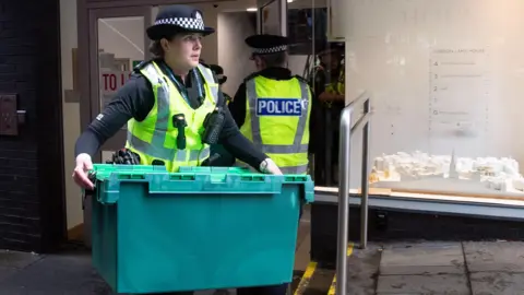 PA Media A female police officer carries a large green plastic box out of the SNP's headquarters in Edinburgh following the arrest of Peter Murrell in April 2023. Another officer is heading back into the building, which has a frosted glass exterior.