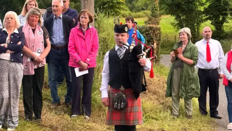 A group of people standing on a grass verge beside a rural road. In front of them is a piper in a red and green plaid kilt.