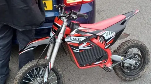 Merseyside Police Red-and-white Neon scrambler bike seized by police resting on the boot of a navy blue car with a person wearing navy trousers and coat standing nearby.