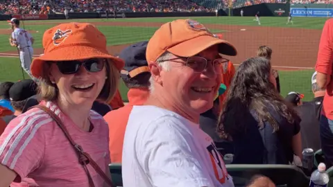 Kristy Gammon Kristy Gammon and her husband at a Baltimore Orioles game. They are both wearing orange Orioles hats and smiling. Behind them is a baseball pitch and some of the crowd. 