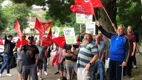 BBC Seveal men holding red Unite flags and placards saying 'Stingy Suez Fair Pay Now'