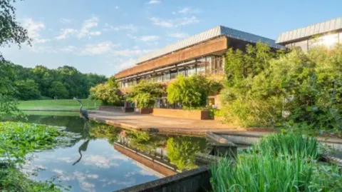 View taken of the HQ building with a pond in front of the building. There are vegetation and trees surrounding the site. The building itself is made up from a mix of concrete and glass and is at least two storeys. 