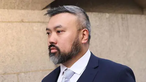 Getty Images A shot of Chi Leung “Peter” Wai from the shoulders up. He has black and grey hair and a black and grey beard. He is wearing a navy suit with a white shirt and a light blue tie. He is walking and looking to the left.