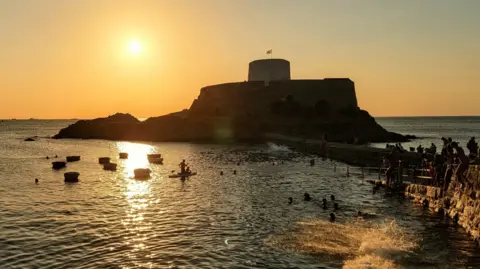 bauzeld Sunset over a coastal harbour with a castle, boats on the water and people swimming and gathering along the shoreline.