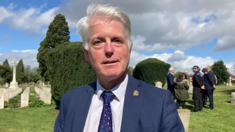 Paul wearing a white shirt and blue blazer. He has short grey hair and is standing in a cemetery and smiling at the camera.