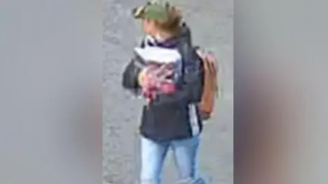 North Yorkshire Police A low quality image of a woman wearing jeans, a dark coat, and a green hat. She is wearing a bag and holding items