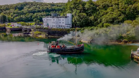 Samuel Callen The Clyde Puffer S.L. VIC 32 - a small steamboat in dark blue/black, with a yellow mast, sails near to the shore. A large white building, many trees and greenspace, and a small number of homes can be seen there. 