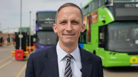 Mr Marion has short light brown hair, and wears a blue suit and tie. Two buses are behind him.