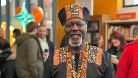 Claire Hamilton/BBC Poet Levi Tafari, who is wearing a black traditional African style gown and hat with an orange pattern, and a necklace with chunky black and orange beads, smiles at the camera