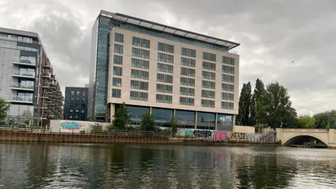 Emma Baugh/BBC A six-storey building with beige cladding and glass sides is pictured from across a river