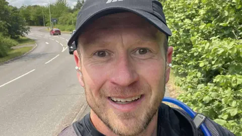 Mike Jones Mike Jones taking a selfie alongside a road with shrubs to the right and the road on the left.