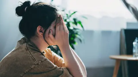 A teenage girl wearing a faded black denim jacket has her head in her hands as she sits on the floor of a room. She has dark hair tied up in a bun. In the background a plant and a small table are visible