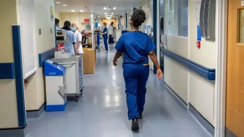 A nurse, wearing blue scrubs, walking through a corridor. There are other healthcare professionals walking through the corridor. The walls are white and blue. There is a bin to the left of the woman and to the side of the bin going up the corridor is a cabinet. 