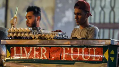 Getty Images Two men make street food behind a cart saying "Liver Fuul" in red.