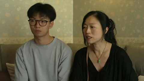 A teenage boy sits on a sofa next to his mother. He is wearing black glasses and a grey jumper. She has her hair tied back and wears a black shirt and a green pendant.