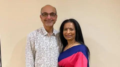 Unknown Kishor Modha with very short dark hair and grey beard with black-framed glasses wearing a black and white shirt. Raxa Modha standing next to him with long dark hair, wearing a pink dress with a blue upper section. They are both smiling at the camera against a cream-coloured background.